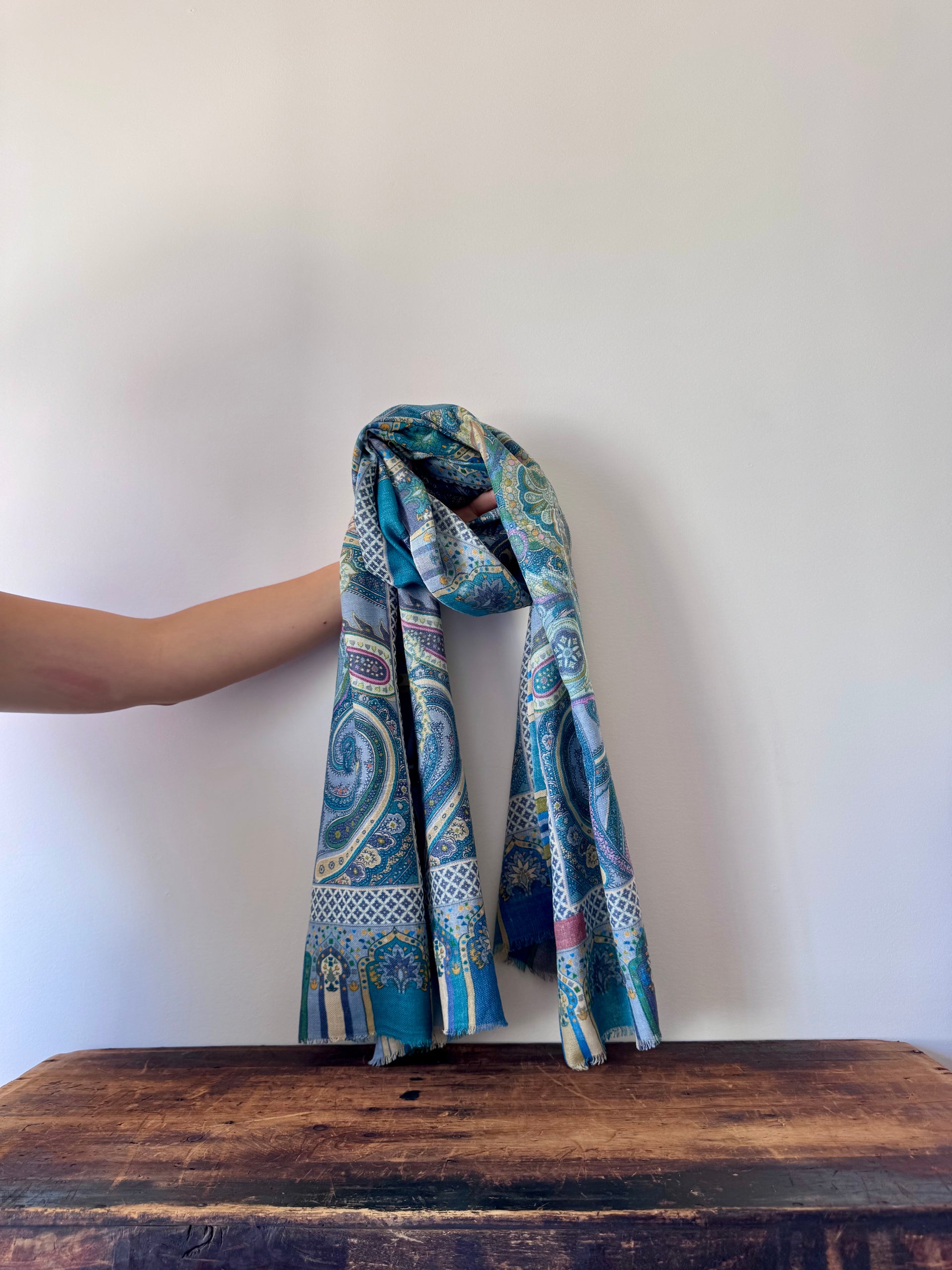 Blue patterned scarf held by a hand on a wooden surface with a white background