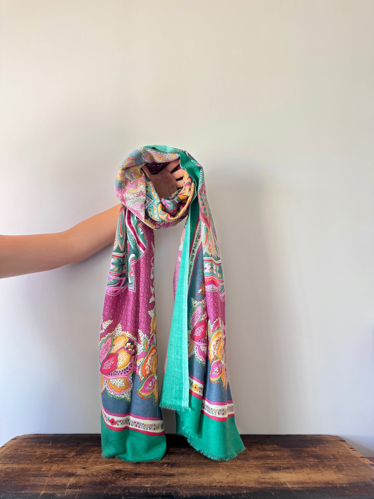 Colorful patterned scarf held by a hand on a wooden surface with a white background