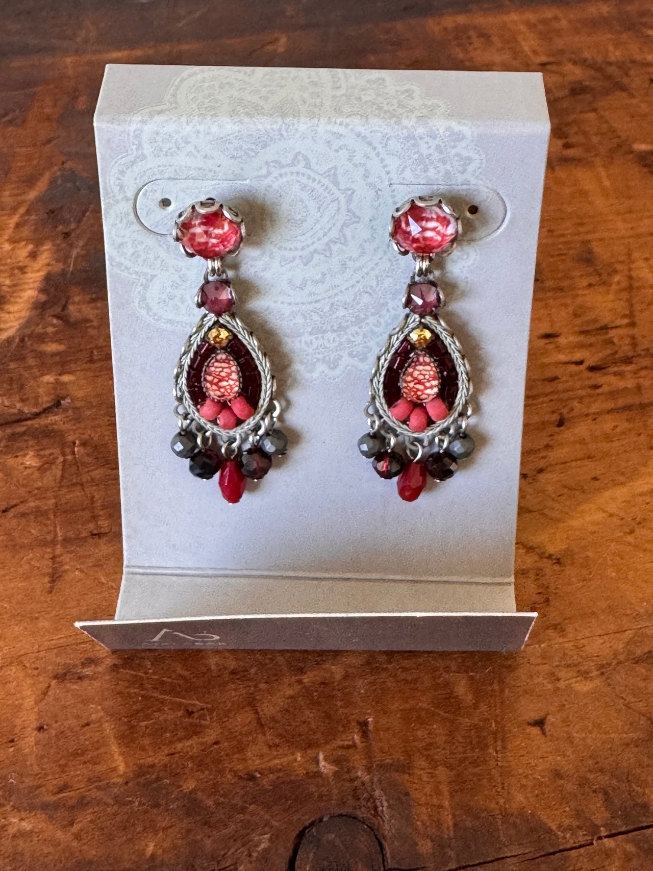 Pair of ornate earrings with red and black beads in a white box on a wooden surface